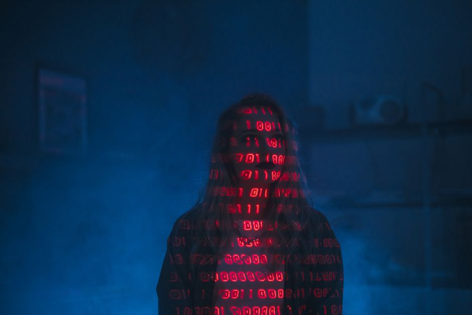 A person standing in a dimly lit environment with their face and upper body illuminated by bright red numbers and code projected onto their dark hoodie, creating a digital or cyber theme. The backgrou