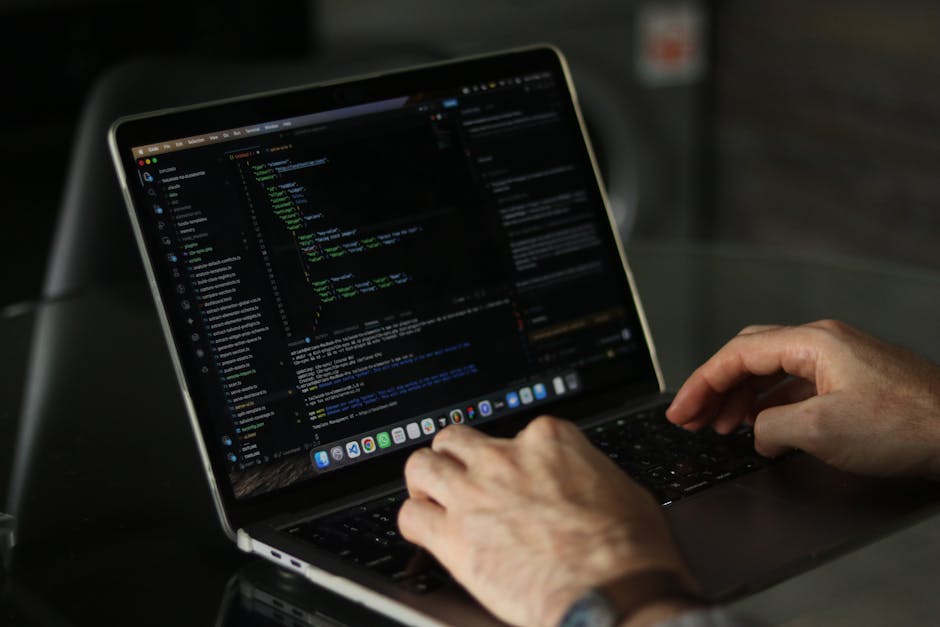 The image shows a person working outdoors, using a laptop placed on a dark surface. The laptop screen displays a dark-themed code editor with multicolored lines of code, indicating active programming 